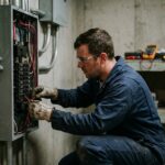 Électricien professionnel en tenue de travail bleue connectant des fils dans un tableau électrique résidentiel, ambiance de chantier réaliste